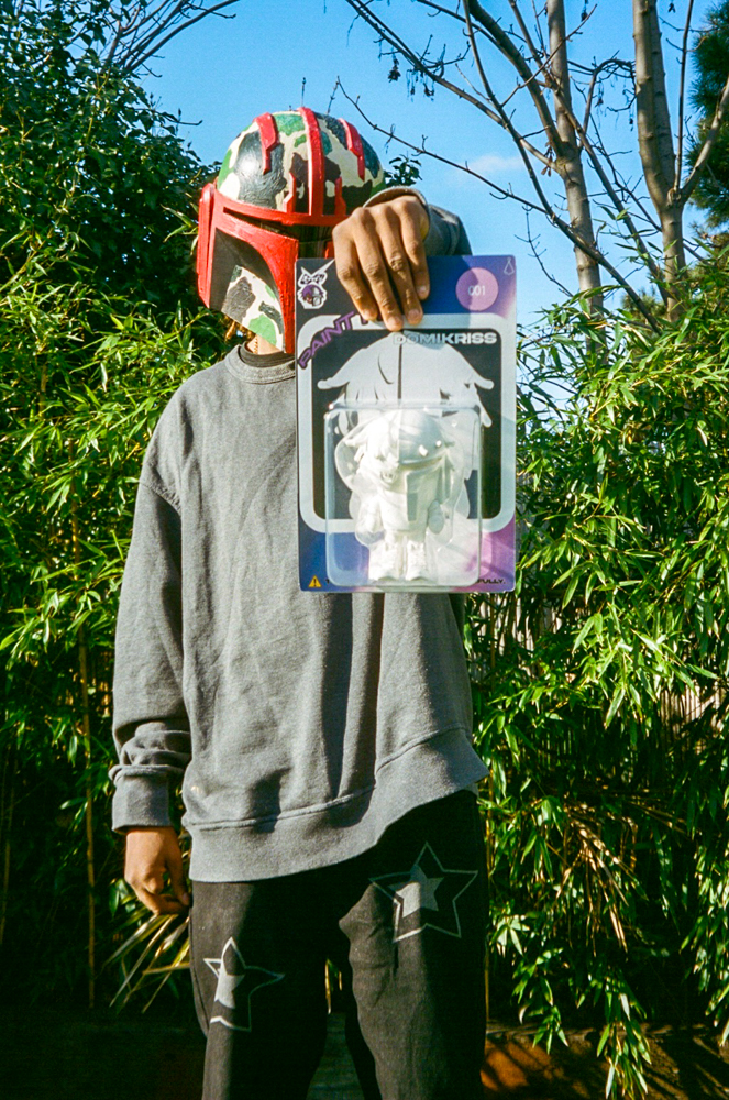 DomiKriss standing, holding 3D printed design object in blister pack, wearing colorfully designed helmet